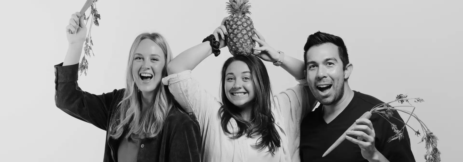Three people smiling and posing playfully; the person in the center balances a pineapple on their head, while the two on each side hold carrots with leafy tops—a lively black-and-white shot perfect for creative healthcare marketing campaigns.