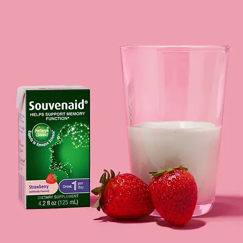A glass of milk sits next to two fresh strawberries and a box of Souvenaid strawberry drink supplement, illustrating consumer health marketing against a pink background.