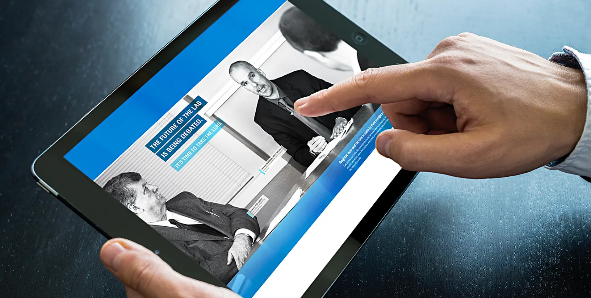 A person uses a tablet displaying a webpage with a black-and-white photo of two men in suits discussing market shaping strategies. One man points at the screen, and the page features blue and white accents with text.