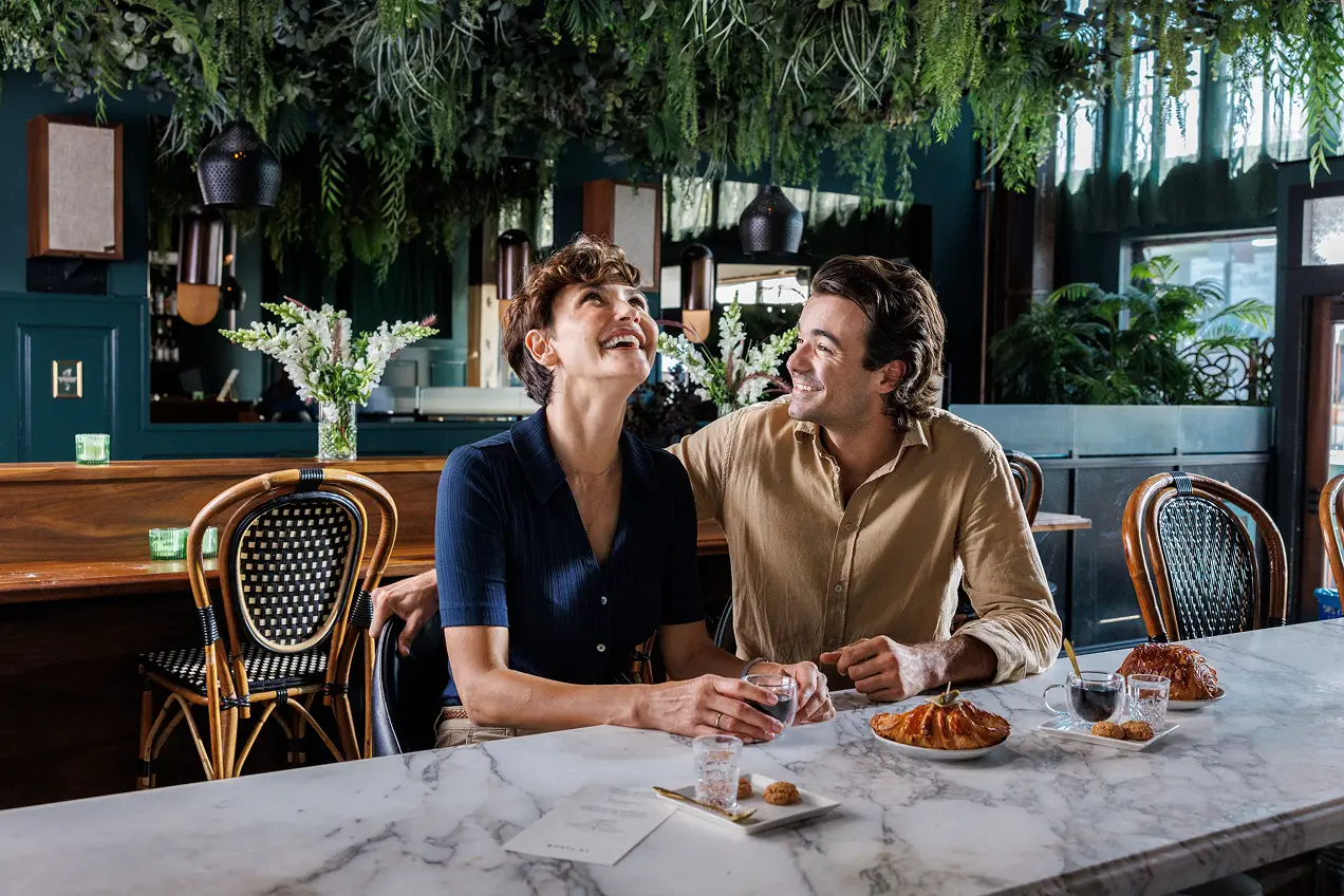 A smiling couple sits at a marble table in a stylish café with greenery overhead, holding hands and enjoying pastries and drinks, their cozy, elegant atmosphere reflecting the thoughtful touches of brand strategy in consumer health marketing.