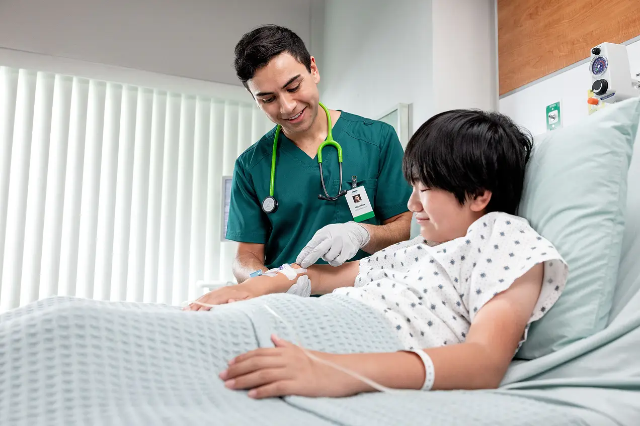A nurse in green scrubs and gloves cares for a smiling young patient in a hospital bed, gently checking the patient’s IV—a compassionate moment that highlights the heart of healthcare marketing. Bright light streams through vertical blinds in the background.