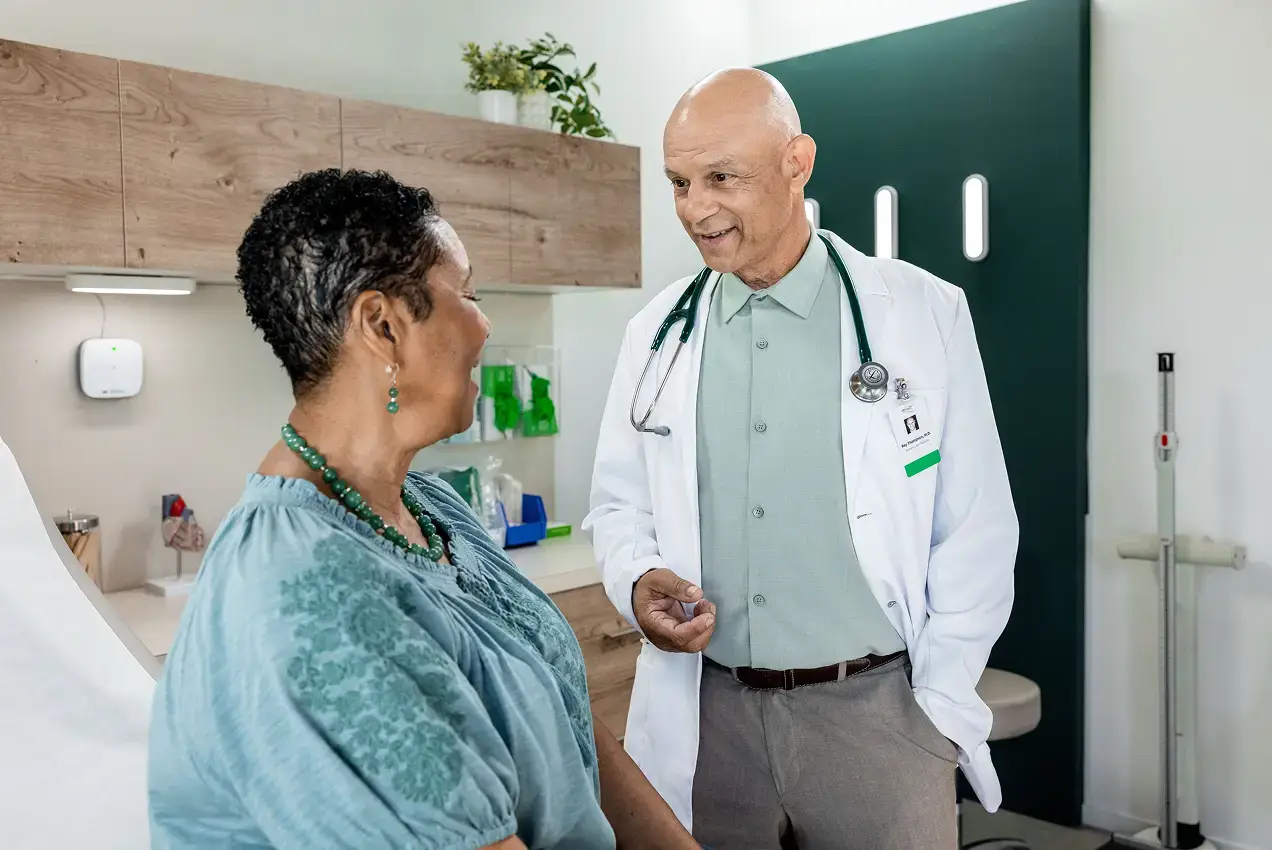 A doctor in a white coat with a stethoscope talks with a smiling patient sitting on an exam table in a modern medical office, illustrating the importance of strong brand strategy in healthcare marketing.