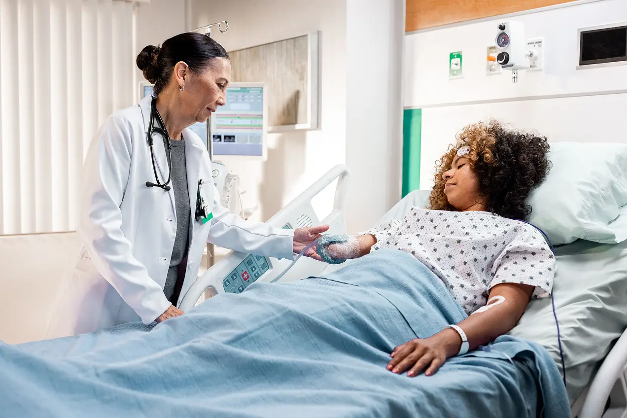 A doctor in a white coat stands beside a hospital bed, gently holding the hand of a patient under a blue blanket. Medical equipment—essential tools in pharma marketing—are visible as the patient rests, IV in arm.