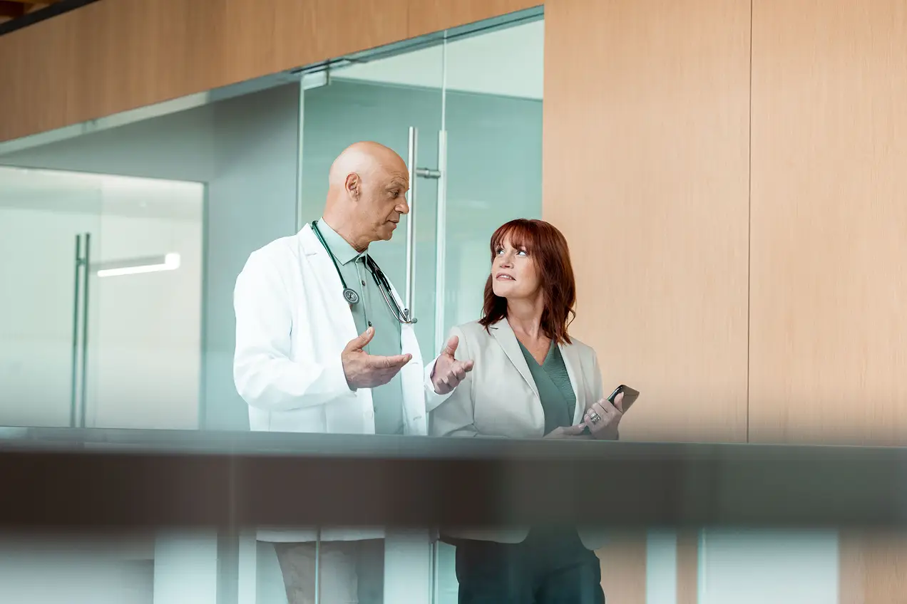 A male doctor in a white coat talks to a woman holding a smartphone as they walk together in a modern office hallway, discussing healthcare marketing strategies amid wooden walls and glass panels.