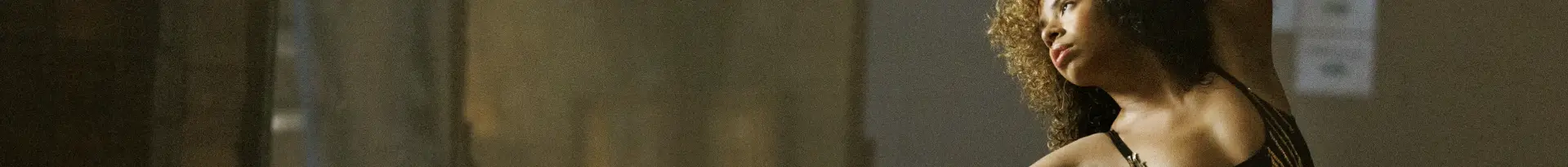 A woman with curly hair and a serious expression stands indoors, wearing a black, strappy top, with a blurred neutral-colored background behind her—capturing the focus needed in fast-paced healthcare marketing.