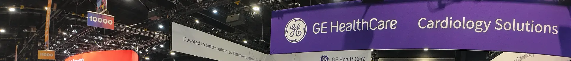 A purple banner at an event displays the GE HealthCare logo and the words Cardiology Solutions, showcasing effective healthcare marketing with other booths and hanging signs visible in the background.