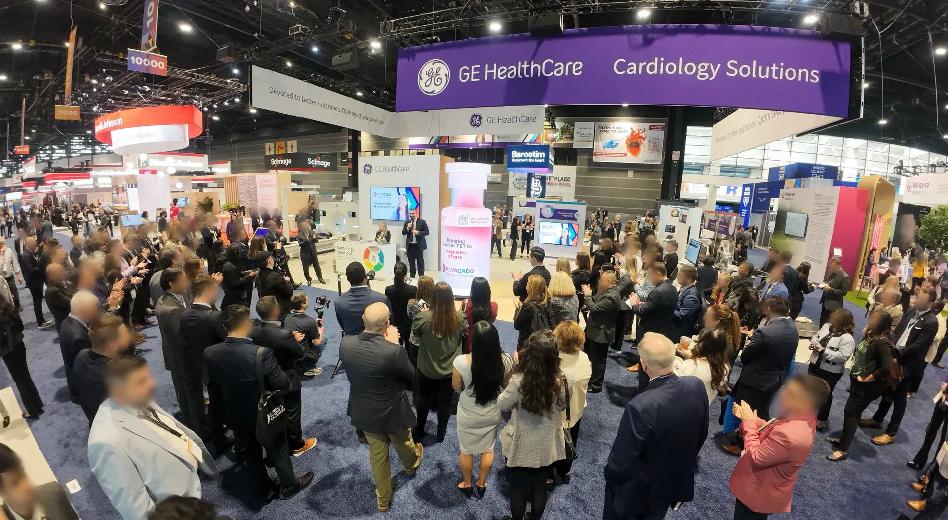 A large crowd gathers at the GE HealthCare Cardiology Solutions booth, where excitement builds around a new cardiac PET radiotracer launch. Attendees face the stage, surrounded by displays and banners in the spacious, well-lit exhibition hall.