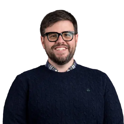 A bearded man with glasses is smiling, wearing a navy blue sweater over a checkered shirt—an approachable look perfect for a healthcare marketing agency. The background is transparent.