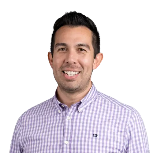 A man with short dark hair smiles at the camera, wearing a light purple and white checkered button-up shirt, standing against a plain white background—a true representative of our healthcare marketing agency’s core values.