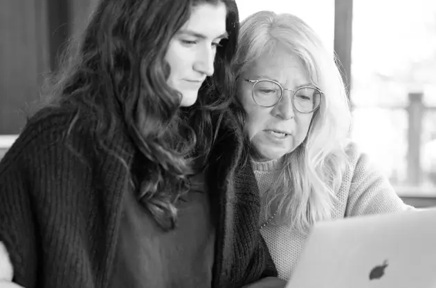 Two women sit closely together, looking at a laptop screen. One has long dark hair and the other has long light hair and glasses. They appear focused, possibly discussing healthcare marketing or strategies for a successful product launch.
