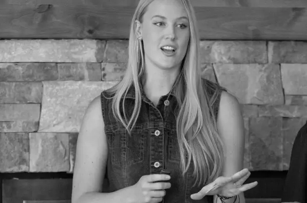 A woman with long blonde hair, wearing a sleeveless denim vest, gestures with one hand while speaking about healthcare marketing. She stands in front of a stone wall. The image is in black and white.