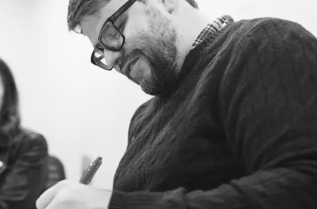 A man wearing glasses and a dark sweater is smiling and writing with a pen on paper. The black-and-white photo captures his focus, suggesting he’s planning a brand strategy or mapping out ideas in healthcare marketing.