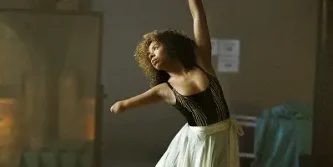 A woman with one arm, wearing a black and gold leotard and a white skirt, dances gracefully with one arm raised in a softly lit studio, embodying the elegance and determination found in successful healthcare marketing.
