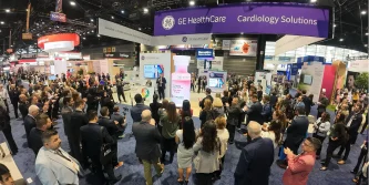 A large crowd gathers at a GE HealthCare Cardiology Solutions booth during a convention, drawn by an engaging medtech marketing presentation under hanging banners in a spacious, well-lit exhibition hall.