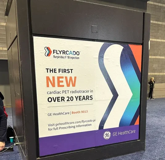 A large display panel highlights a FLYRCADO advertisement, featuring GE HealthCare’s first new cardiac PET radiotracer in 20 years—a product launch that showcases innovative medtech marketing. Booth info and a blue-orange abstract design are included.
