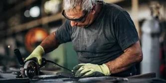 An older man wearing safety glasses and gloves uses a handheld grinder on a metal surface in an industrial workshop, with sparks flying from the tool—demonstrating precision and focus essential in fields like medtech marketing.
